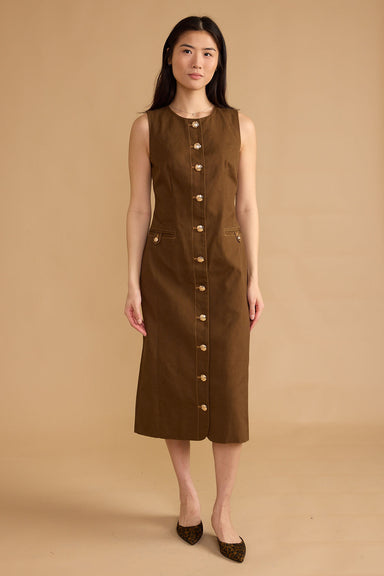 A woman modeling a sleeveless brown midi dress with a button-down front and side pockets, standing against a solid beige studio backdrop.
