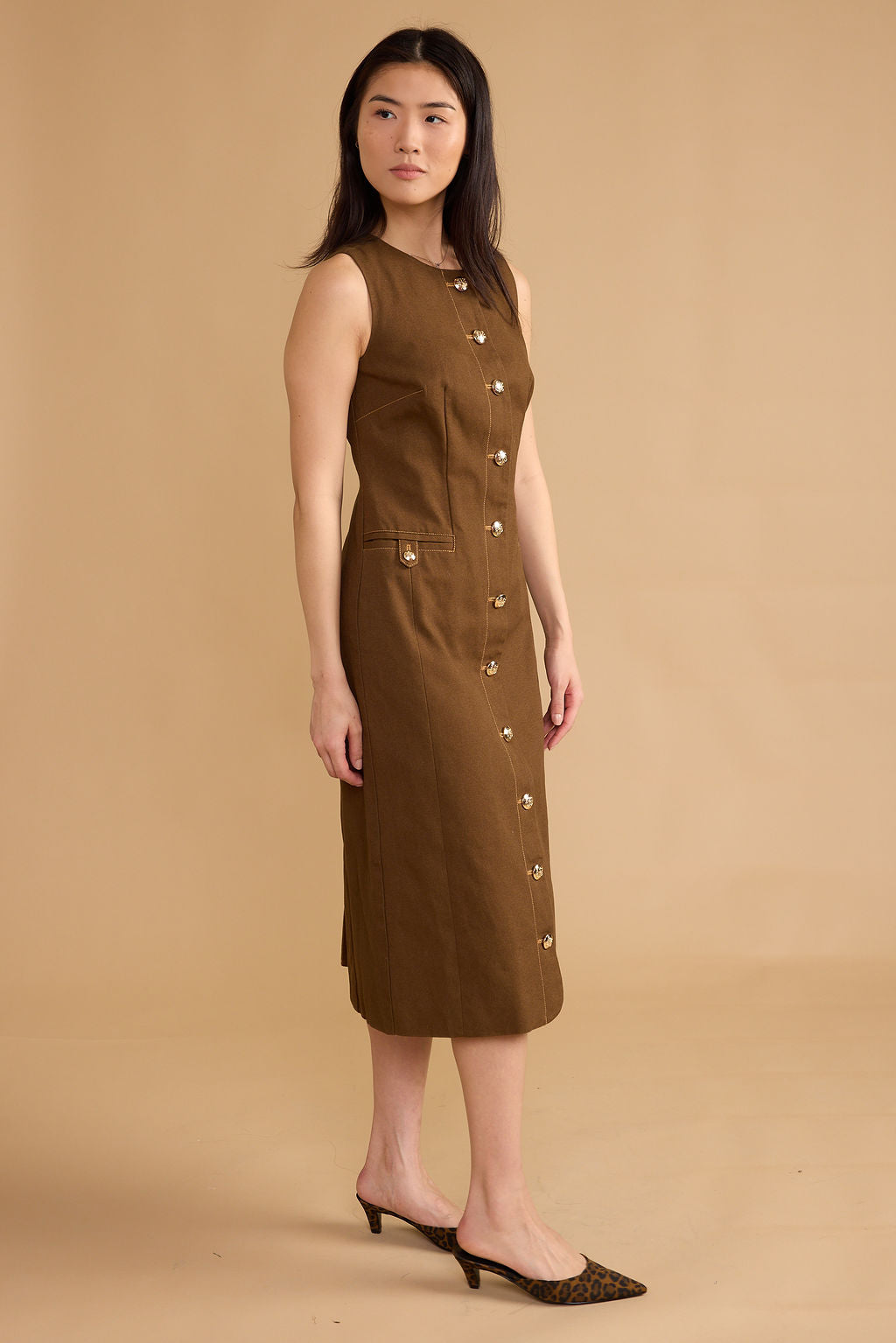 A woman modeling a sleeveless brown midi dress with a front button placket and side pockets, paired with brown pointed-toe mule heels, against a solid beige studio background.