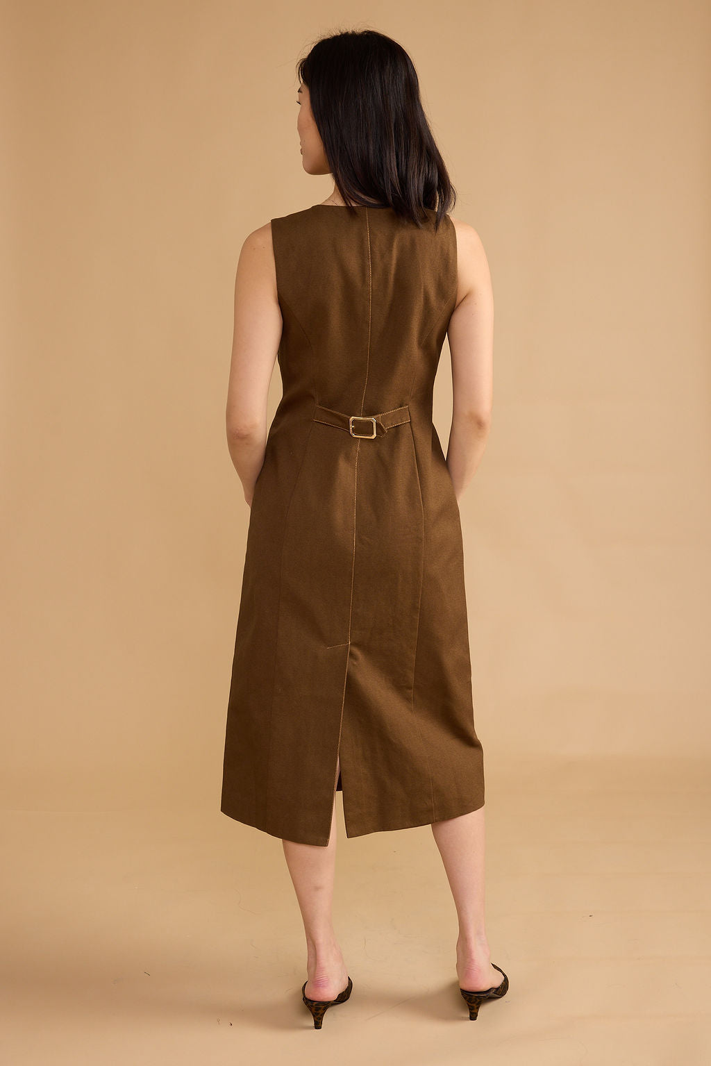 A woman viewed from the back wearing a sleeveless brown midi dress with a matching belt and small gold-tone buckle, standing against a plain beige studio backdrop.