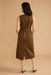 A woman viewed from the back wearing a sleeveless brown midi dress with a matching belt and small gold-tone buckle, standing against a plain beige studio backdrop.