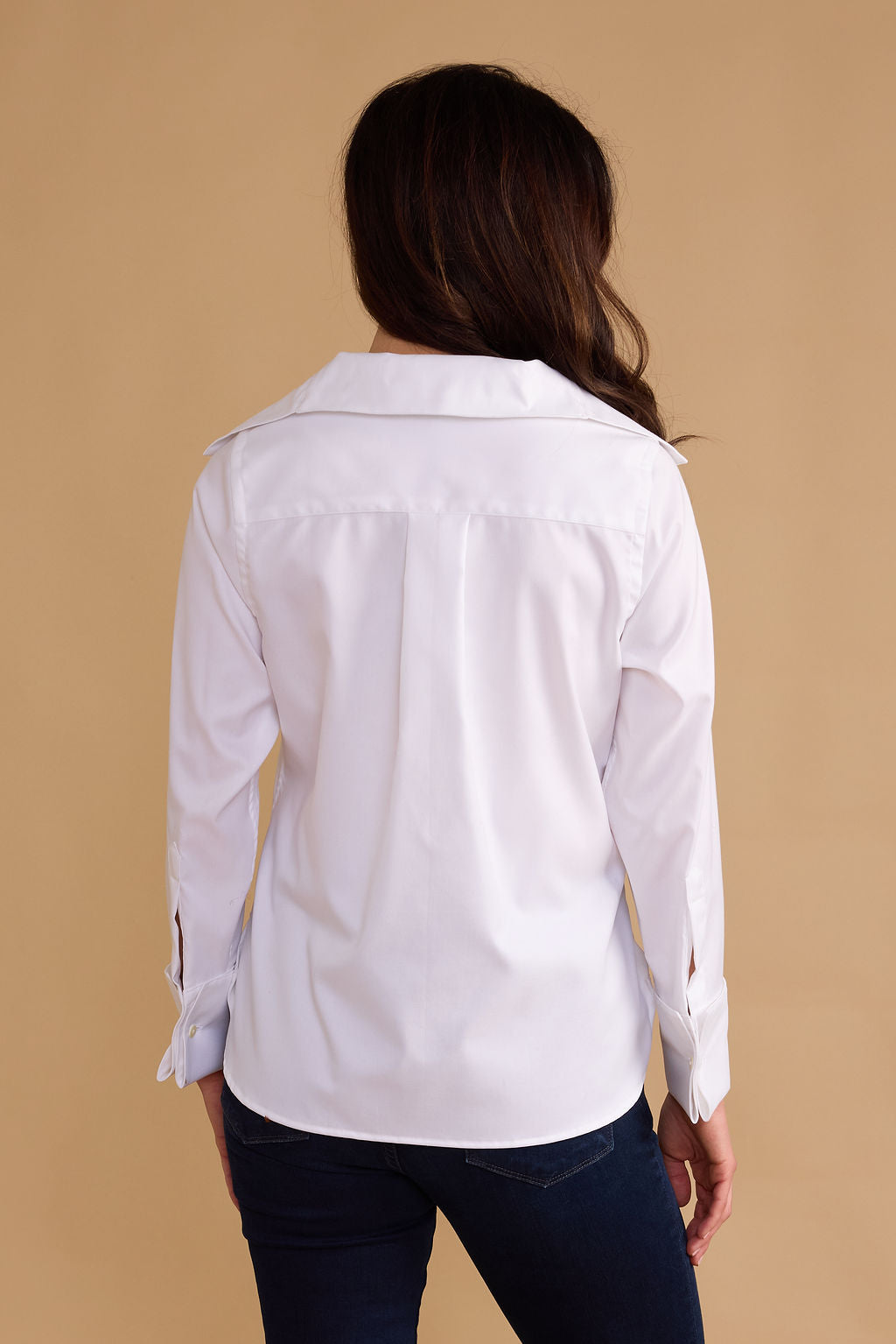 A woman viewed from the back wearing a white long-sleeve button-up shirt and dark blue jeans, standing against a solid beige background.