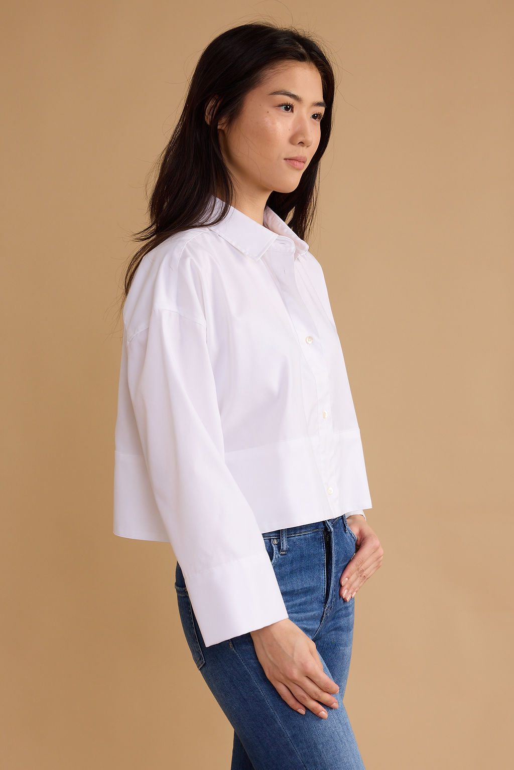 A woman wearing a white long-sleeve button-up shirt and blue jeans, shown in profile against a solid beige studio background.