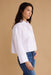 A woman wearing a white long-sleeve button-up shirt and blue jeans, shown in profile against a solid beige studio background.