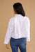 A woman shown from the back wearing a white long-sleeve cropped blouse and blue jeans, standing against a solid warm beige studio background.