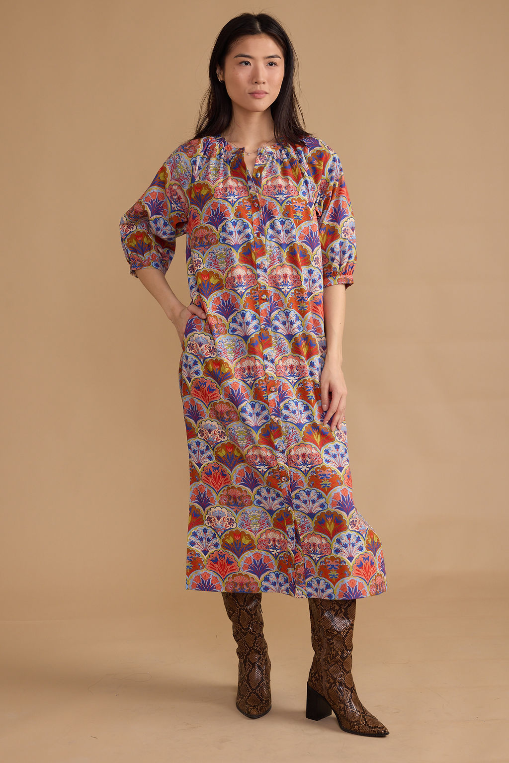 A woman modeling a long-sleeved midi dress with a colorful, dense bohemian fan and floral pattern in red, blue, orange, and white, paired with brown snakeskin-patterned boots against a solid beige studio background.