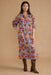 A woman modeling a long-sleeved midi dress with a colorful, dense bohemian fan and floral pattern in red, blue, orange, and white, paired with brown snakeskin-patterned boots against a solid beige studio background.