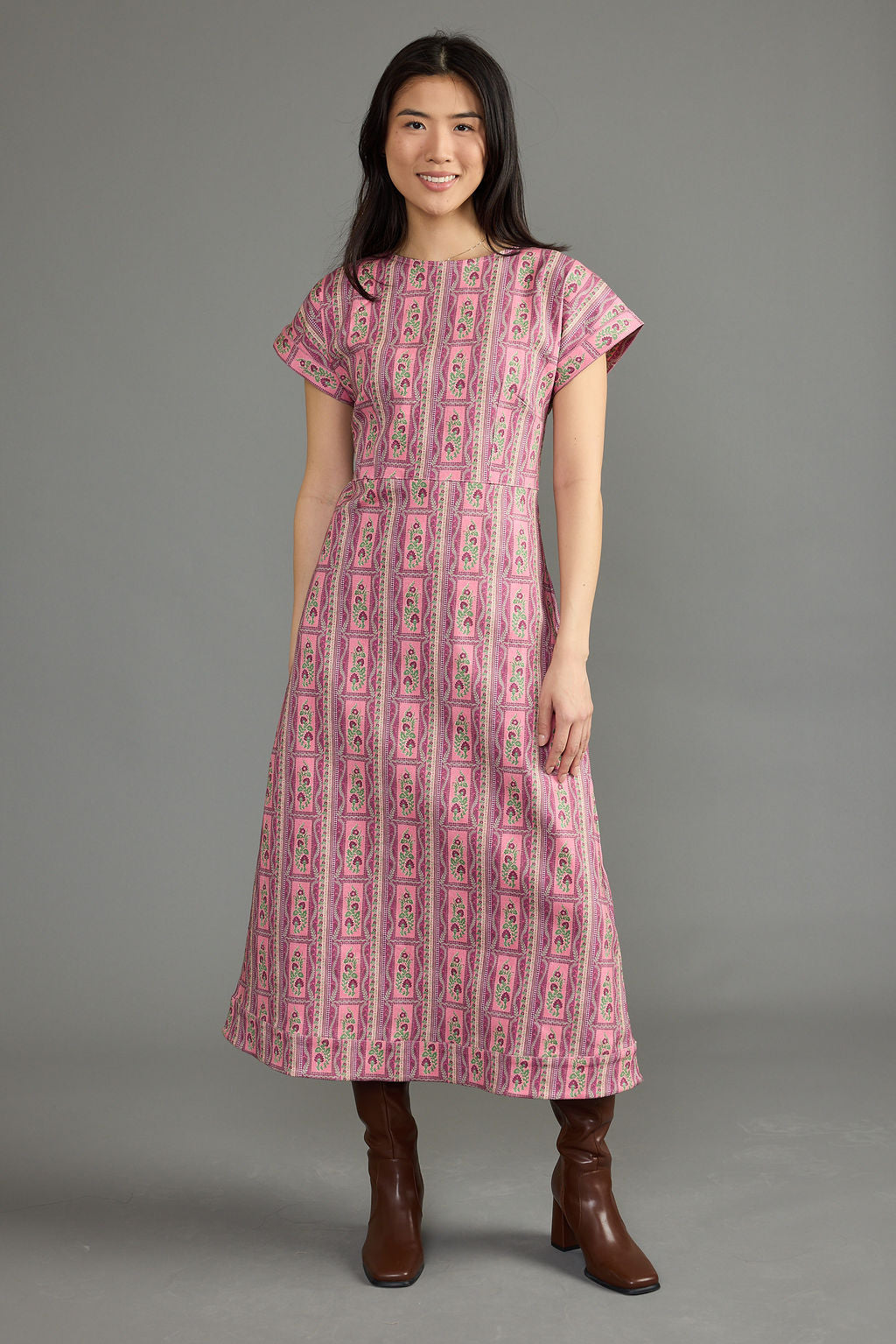 A woman modeling a pink midi dress with a vertical multicolor geometric pattern, paired with brown knee-high boots, against a solid gray studio background.