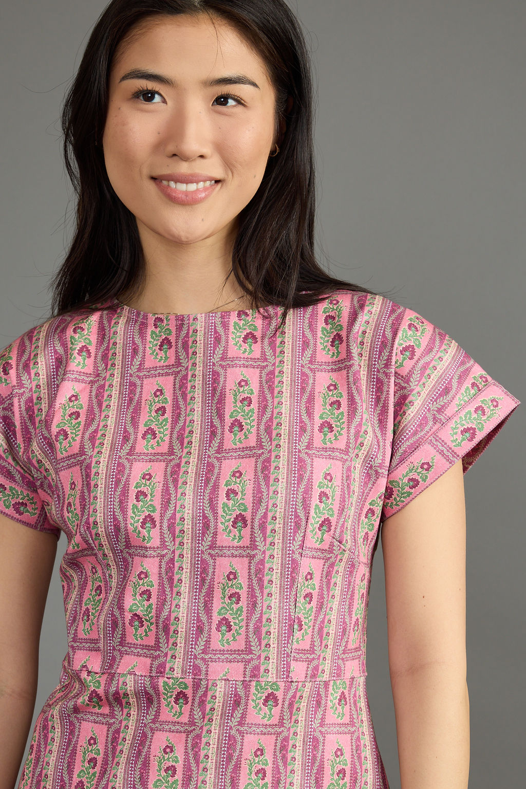 A woman wearing a pink short-sleeve dress with a vertical floral block-print pattern in green, yellow, and white, photographed against a solid gray studio background.