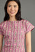 A woman wearing a pink short-sleeve dress with a vertical floral block-print pattern in green, yellow, and white, photographed against a solid gray studio background.