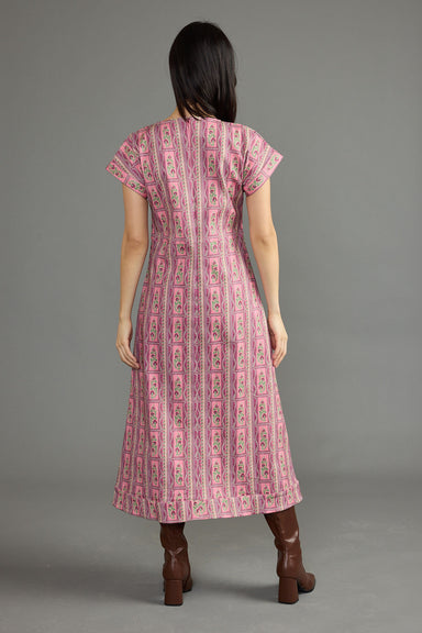 A woman viewed from the back wearing a pink patterned midi dress with short sleeves and brown boots, standing against a plain gray studio background.