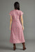 A woman viewed from the back wearing a pink patterned midi dress with short sleeves and brown boots, standing against a plain gray studio background.