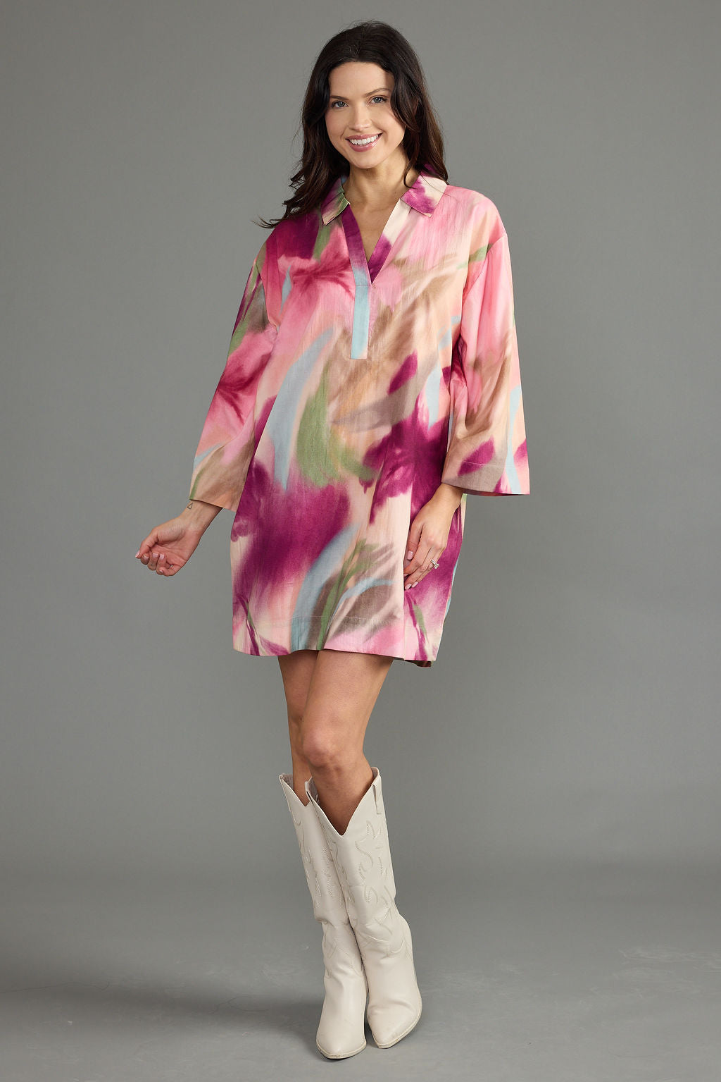 A woman modeling a short, loose-fitting button-front tunic dress with three-quarter sleeves in a pink, peach, and purple abstract print, paired with white cowboy boots against a solid gray studio background.