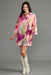 A woman modeling a short, loose-fitting button-front tunic dress with three-quarter sleeves in a pink, peach, and purple abstract print, paired with white cowboy boots against a solid gray studio background.
