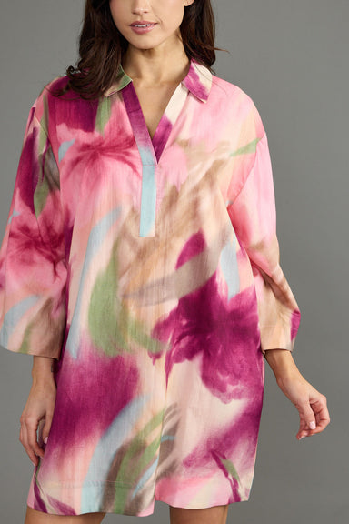 A woman wearing a loose, short-sleeve collared tunic dress with a pink, magenta, beige, and green abstract floral print, photographed against a solid gray studio background.