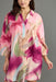 A woman wearing a loose, short-sleeve collared tunic dress with a pink, magenta, beige, and green abstract floral print, photographed against a solid gray studio background.