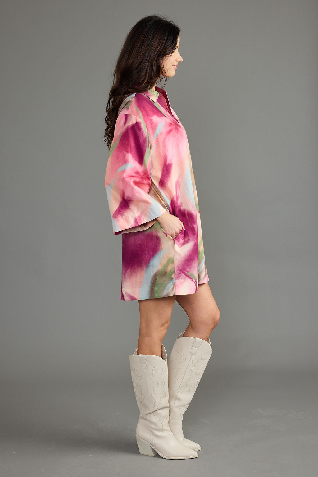 A woman in profile wearing a short pink and purple floral print coat with wide sleeves, paired with off-white knee-high boots, against a solid gray studio background.