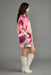 A woman in profile wearing a short pink and purple floral print coat with wide sleeves, paired with off-white knee-high boots, against a solid gray studio background.