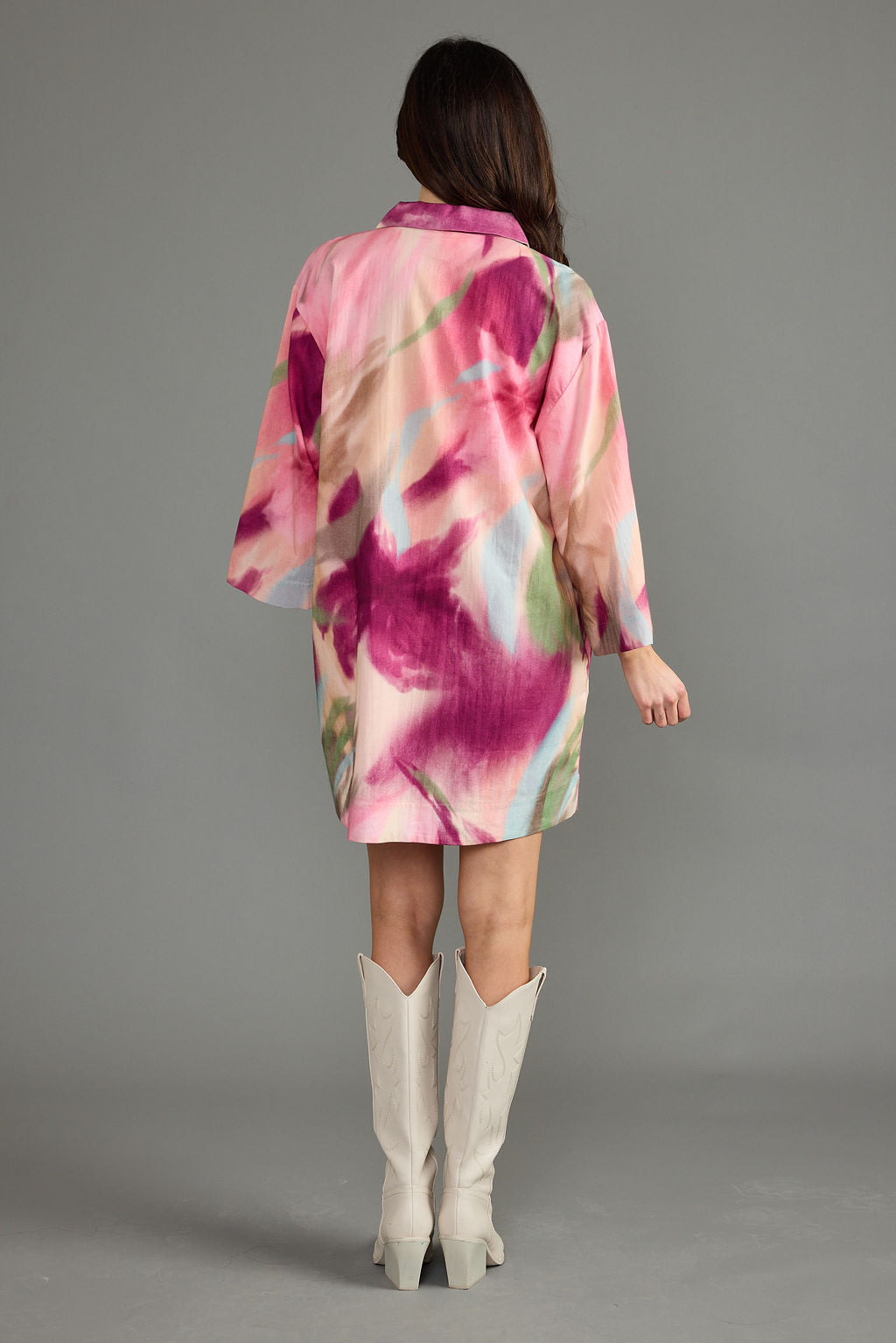 A woman viewed from the back wearing a short pink kimono-style robe with a large watercolor floral print in magenta, peach, and green, paired with off-white cowboy boots against a solid gray studio background.