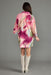 A woman viewed from the back wearing a short pink kimono-style robe with a large watercolor floral print in magenta, peach, and green, paired with off-white cowboy boots against a solid gray studio background.