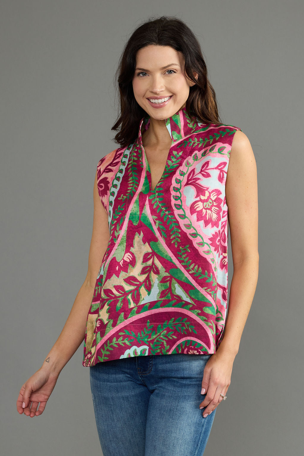 A woman modeling a sleeveless wrap-style top with a vibrant paisley and floral pattern in pink, green, and beige, paired with blue jeans against a solid gray studio background.
