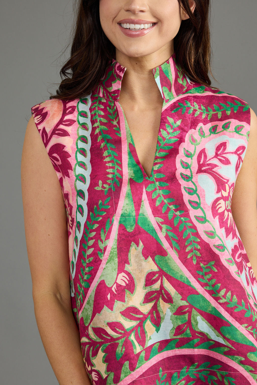 A close-up of a woman wearing a sleeveless high-neck top with a vibrant pink, green, and beige botanical print against a solid gray background.