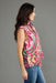 A woman shown in side profile wearing a sleeveless collared blouse with a bold pink, green, and beige paisley-style print, paired with blue jeans, against a solid gray studio background.