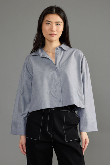 A woman modeling a light blue cropped button-up shirt with a relaxed fit and black pants against a solid gray studio background.