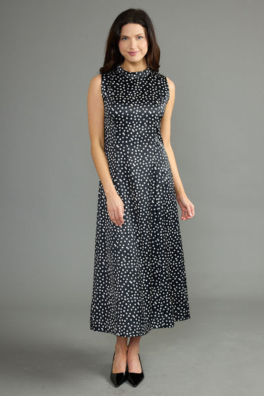 A woman modeling a sleeveless black midi dress with a white polka dot pattern, standing against a solid gray studio background.