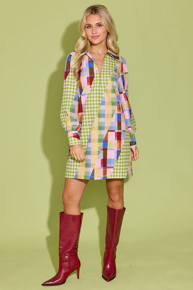 A woman modeling a long-sleeve mini shirt dress with a colorful patchwork gingham pattern, paired with shiny burgundy knee-high boots, against a solid light green background.