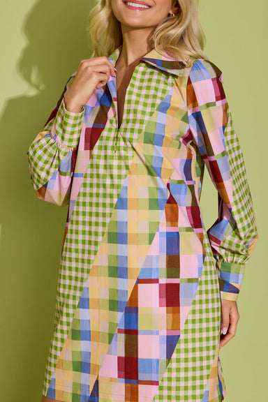 A woman wearing a long-sleeve collared dress with a colorful geometric patchwork print and green gingham sections, posed against a solid olive green background.