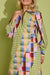 A woman wearing a long-sleeve collared dress with a colorful geometric patchwork print and green gingham sections, posed against a solid olive green background.