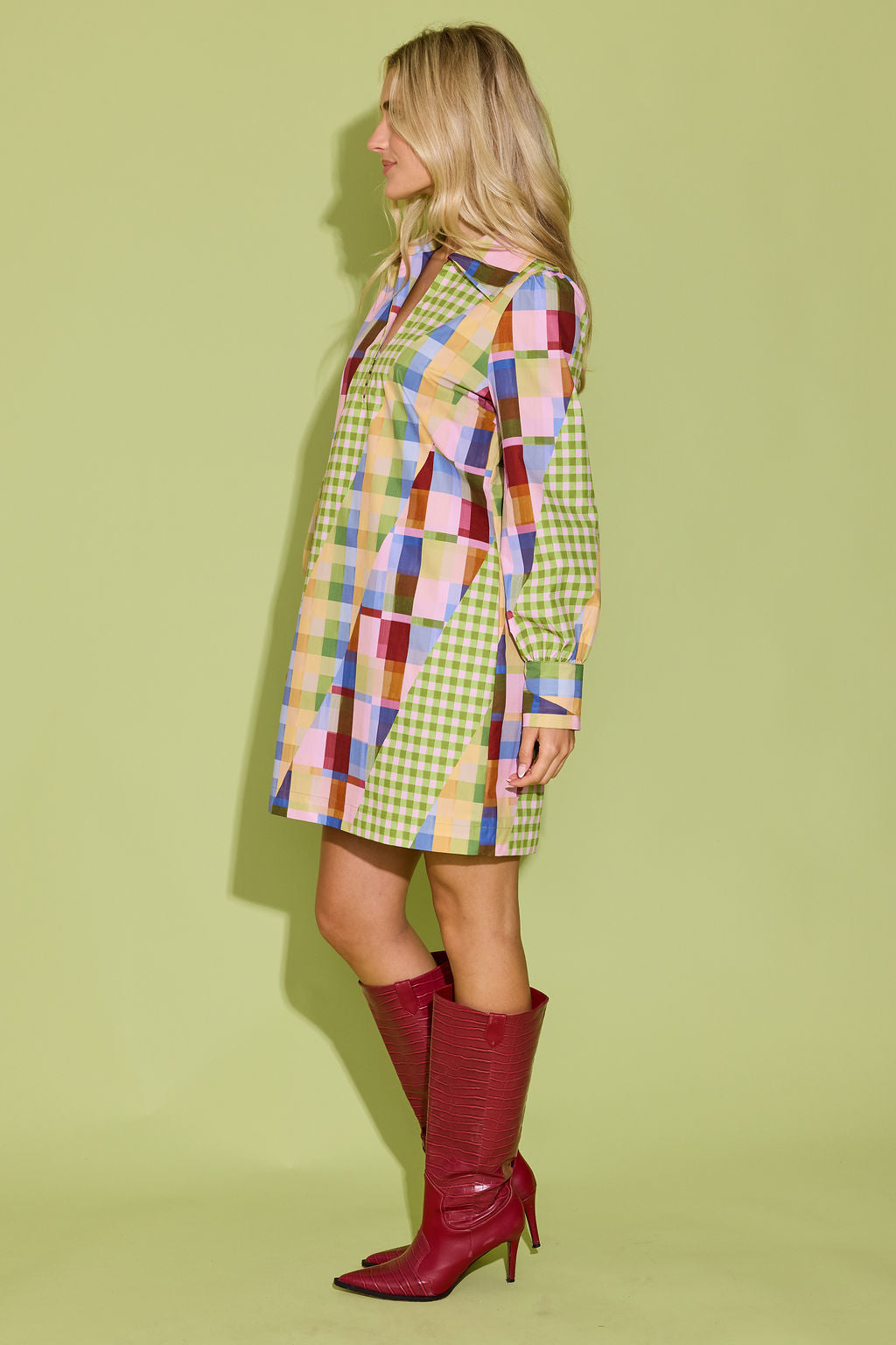 A woman stands in profile wearing a long-sleeve mini shirt dress with a colorful patchwork geometric print and red knee-high heeled boots, set against a solid light green studio background.