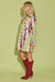 A woman stands in profile wearing a long-sleeve mini shirt dress with a colorful patchwork geometric print and red knee-high heeled boots, set against a solid light green studio background.