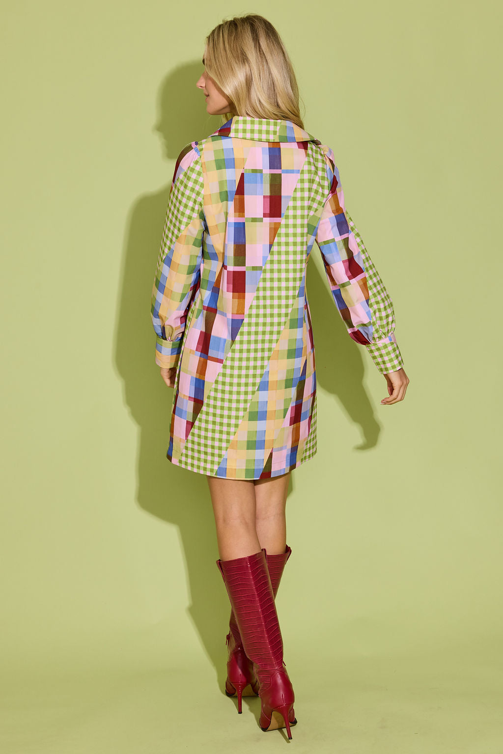 A woman shown from the back wearing a long-sleeve patchwork plaid mini coat in green, yellow, pink, blue, and red, paired with red knee-high boots, against a solid light green studio background.