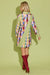 A woman shown from the back wearing a long-sleeve patchwork plaid mini coat in green, yellow, pink, blue, and red, paired with red knee-high boots, against a solid light green studio background.
