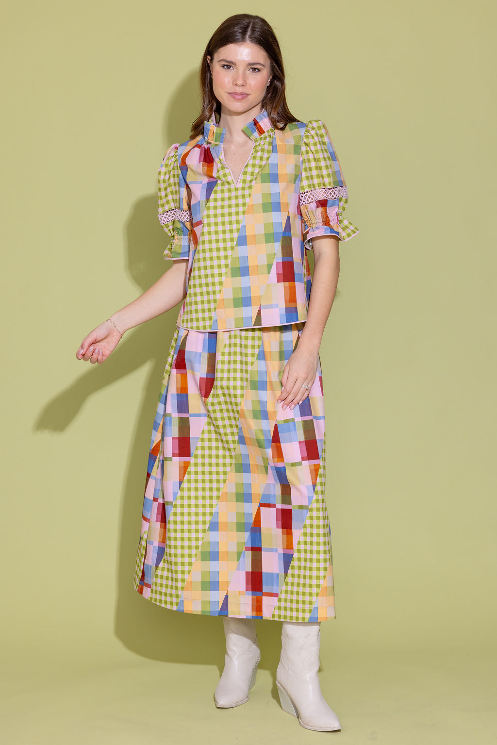 A woman modeling a colorful patchwork and gingham dress with puff sleeves, paired with white ankle boots, against a solid pale yellow-green studio background.