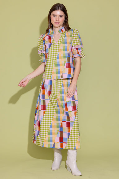 A woman modeling a colorful patchwork and gingham dress with puff sleeves, paired with white ankle boots, against a solid pale yellow-green studio background.