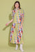 A woman modeling a colorful patchwork and gingham dress with puff sleeves, paired with white ankle boots, against a solid pale yellow-green studio background.
