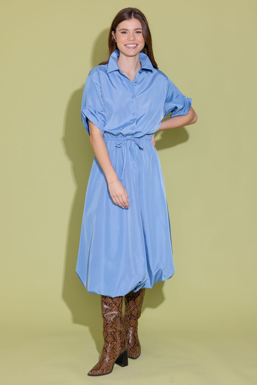 Woman wearing a blue dress with long sleeves and a belt, standing against a light green background.