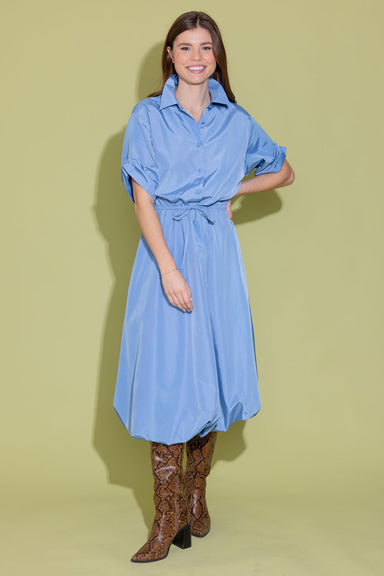 Woman wearing a blue dress with long sleeves and a belt, standing against a light green background.