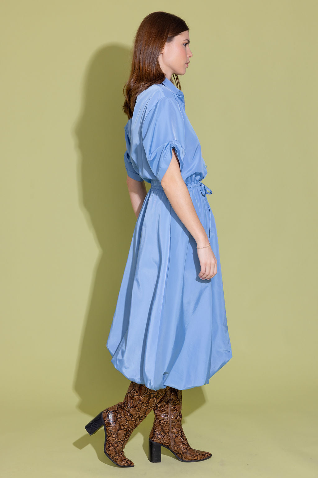 A woman modeling a light blue midi shirt dress with puff sleeves and a tie waist, paired with brown snakeskin-patterned heeled boots, set against a solid olive green studio background.
