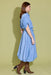 A woman modeling a light blue midi shirt dress with puff sleeves and a tie waist, paired with brown snakeskin-patterned heeled boots, set against a solid olive green studio background.