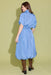 A woman wearing a light blue midi shirt dress with puff sleeves and a gathered waist, shown from the back against a solid pale yellow-green studio background.